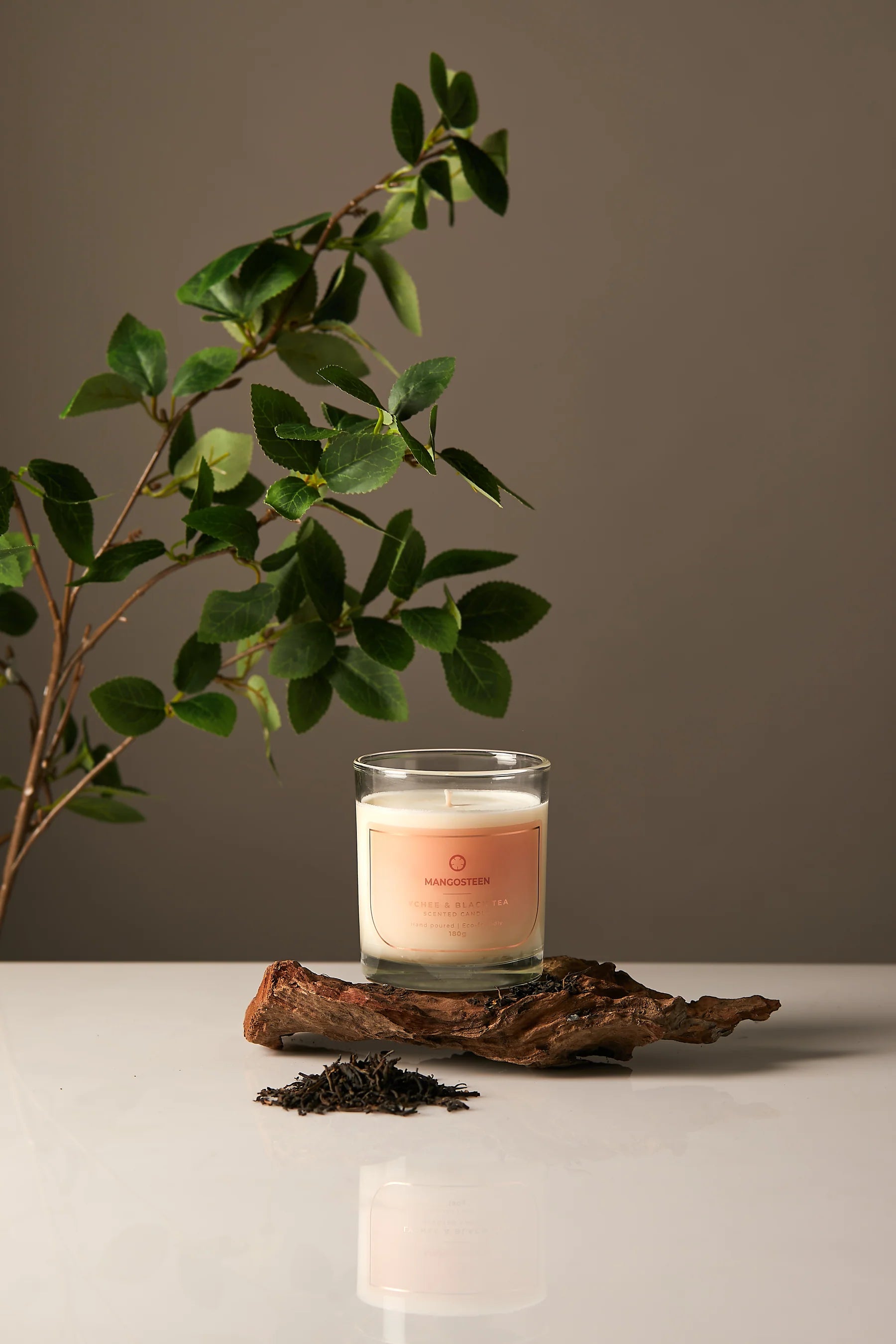 Candle in a glass jar on a wooden log with a plant and tea leaves on a neutral background