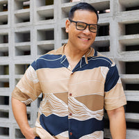 a lifestyle photo of man leaning by wall of brick wearing batik cuban shirt in black bukit pattern