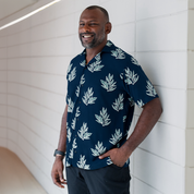 a man wearing a batik shirt in the pattern navy olive leaf in a lifestye photo against a white brick wall