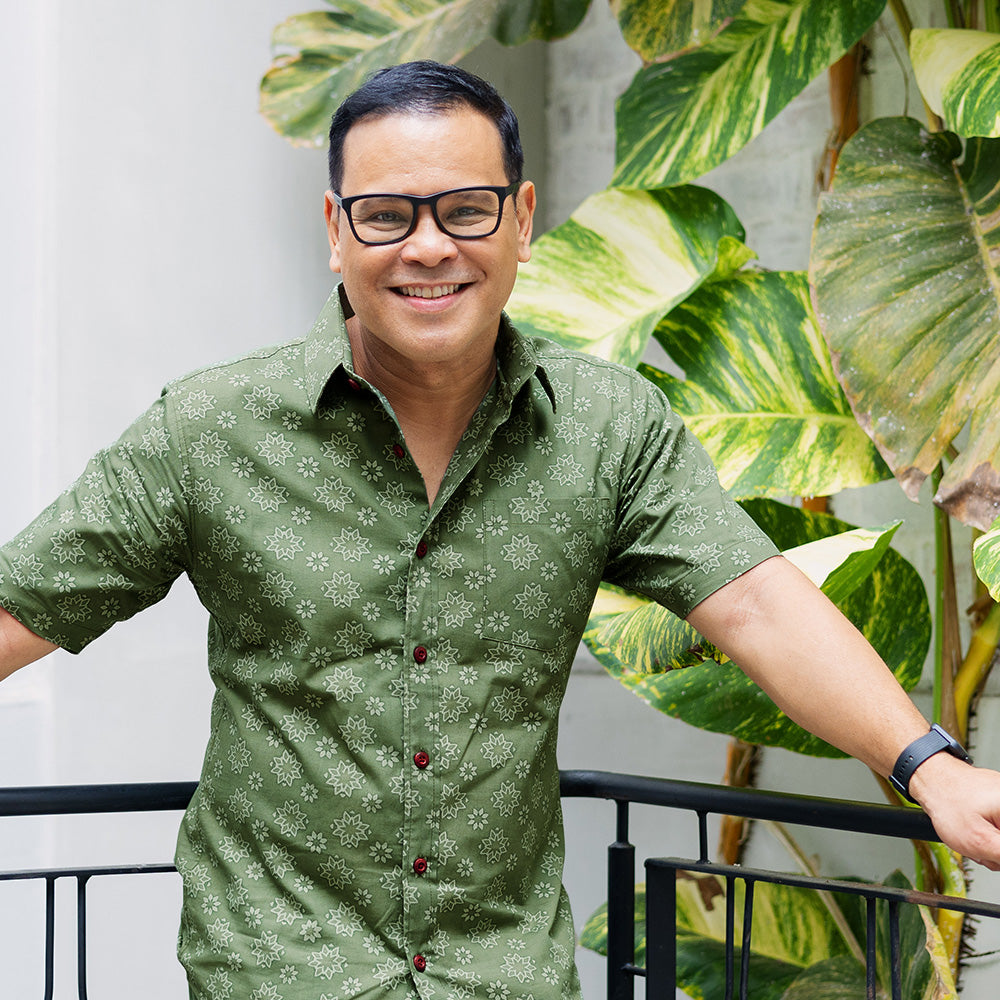 a lifestyle photo of men wearing short sleeve shirt in olive Bintang pattern standing in front of leaf and by the stairs rail