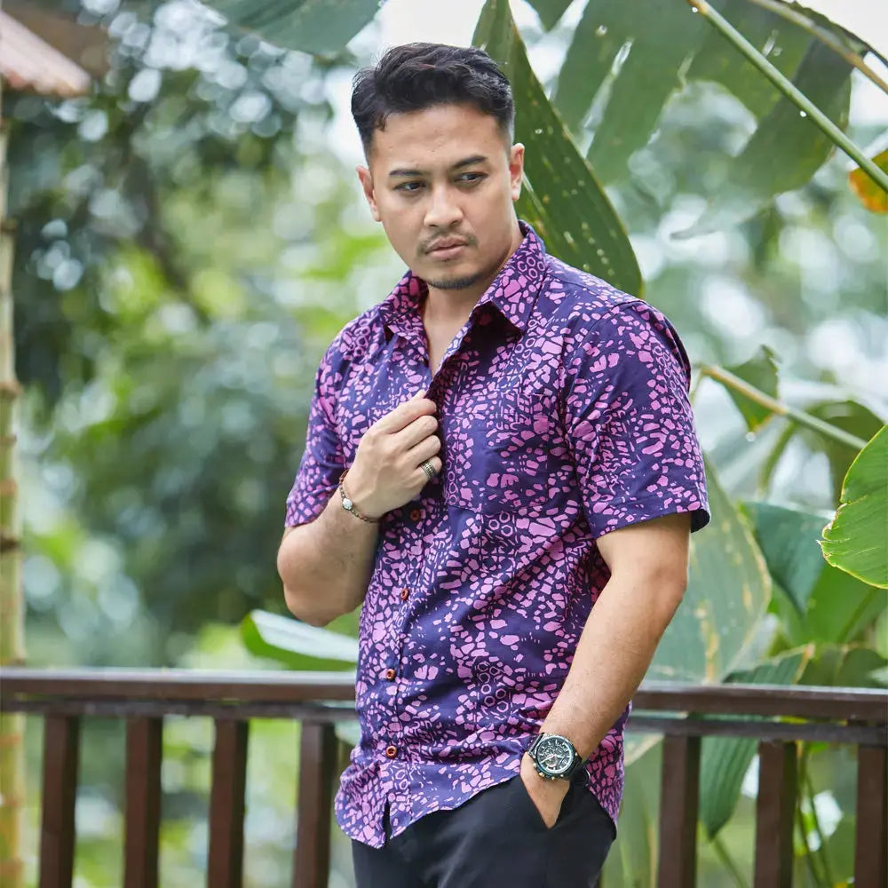 In a picturesque lifestyle photo, a male model strikes a pose amidst lush green surroundings, elegantly dressed in a Purple Bintik patterned batik shirt