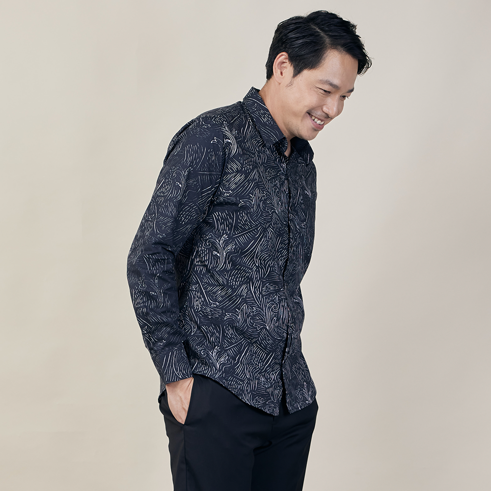 A man striking a pose with a sidelong gaze while elegantly donning an authentic Black Driftwood patterned batik shirt, set against a neutral background