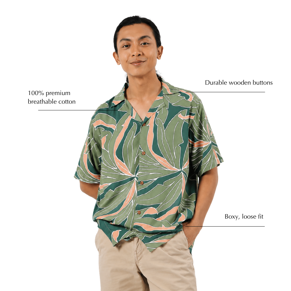 A man wearing a short-sleeve button-up shirt with a green, beige, and pink tropical leaf pattern, paired with khaki pants. Text overlays highlight the shirt's features: '100% premium breathable cotton', 'Durable Wooden buttons', and 'Boxy, loose fit'.