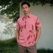 A man wearing a pink short-sleeve button-up shirt with a dark red floral pattern, paired with brown pants, standing outdoors in front of a light-colored wall and green foliage.