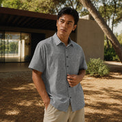 A man wearing a short-sleeve button-up shirt with a fine gray and white micro-pattern, standing outdoors in front of a modern house with trees and dry grass.