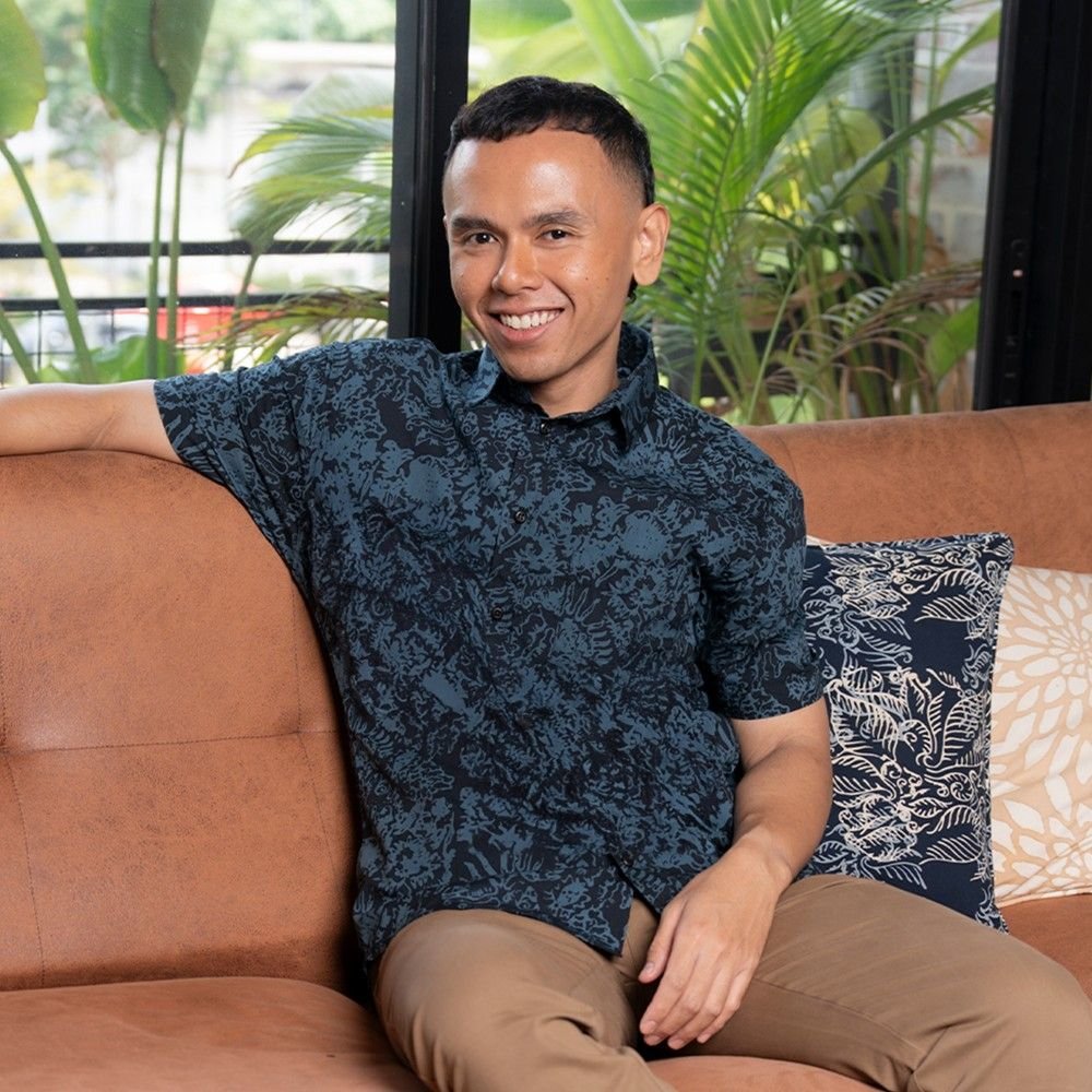 Man sitting on a brown couch wearing a dark patterned shirt with plants in the background