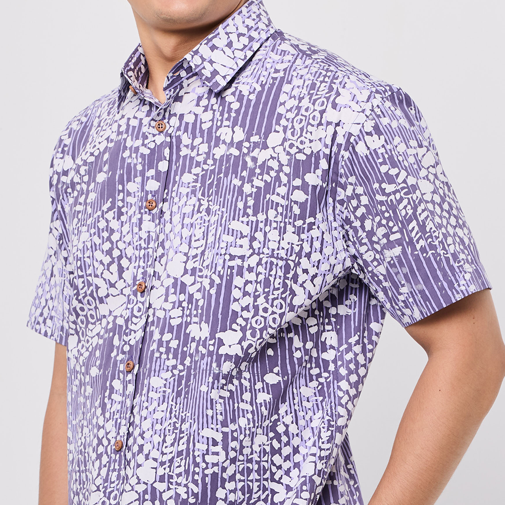 Person wearing a short-sleeve Batik shirt with a purple and white pattern on a plain background