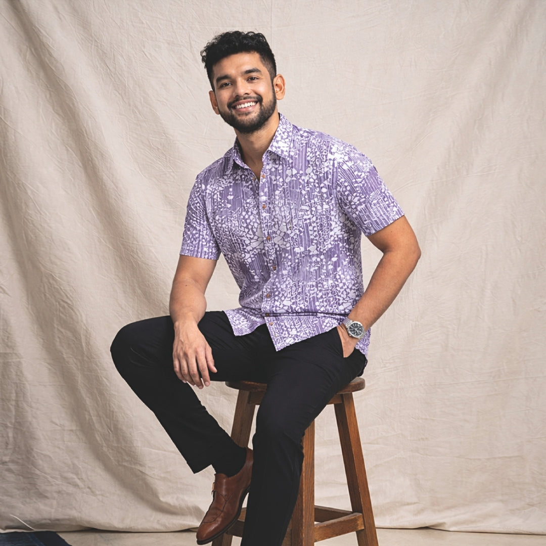 Man sitting on a wooden stool wearing a patterned Batik shirt and black pants against a beige background