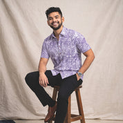 Man sitting on a wooden stool wearing a patterned Batik shirt and black pants against a beige background