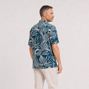 A man wearing a blue button-up shirt with a blue shell pattern, facing his back on a white background.
