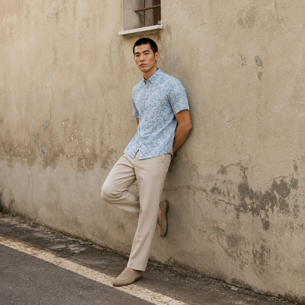 Man leaning against a textured wall wearing a patterned shirt and beige pants.