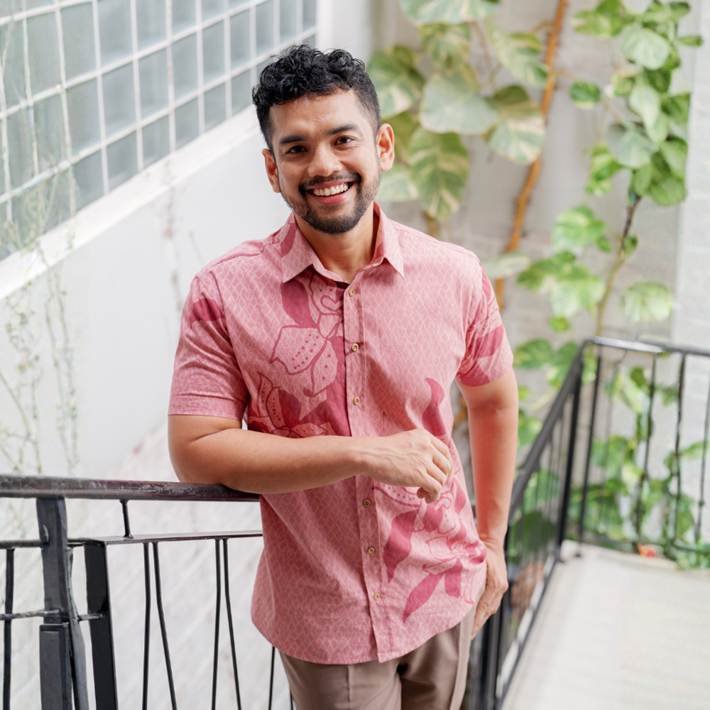Man wearing a pink shirt with leaf patterns standing on a balcony with greenery.
