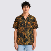 A man modeling a short-sleeve button-up camp shirt with a black and mustard yellow abstract pattern, paired with light blue jeans, against a solid light gray background.