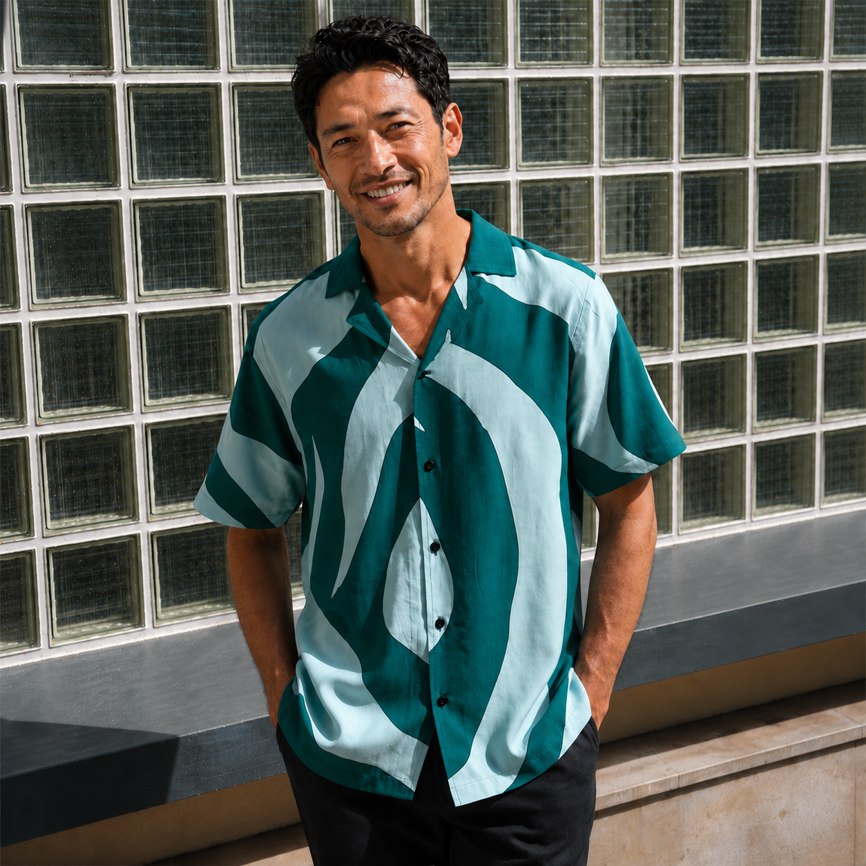 Man wearing green printed batik shirt, with floral patterns posing outside with hands inside his pockets, smiling
