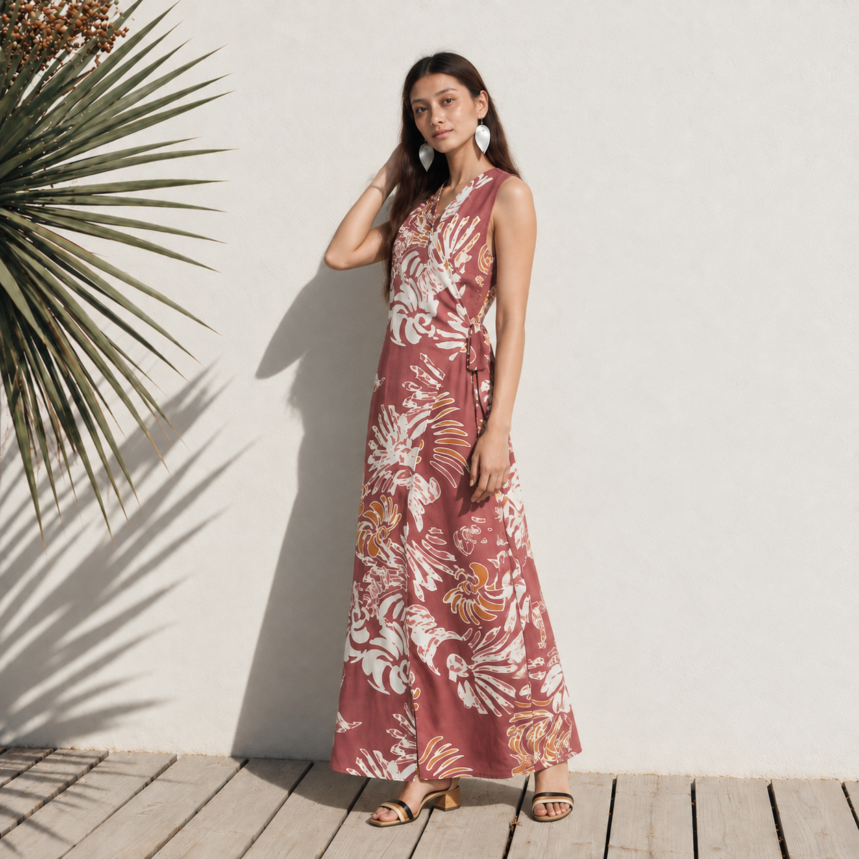 A woman stands on a wooden infront of a white wall wearing a sleeveless, wrap-style maxi dress with a side tie and a light maroon reef pattern.