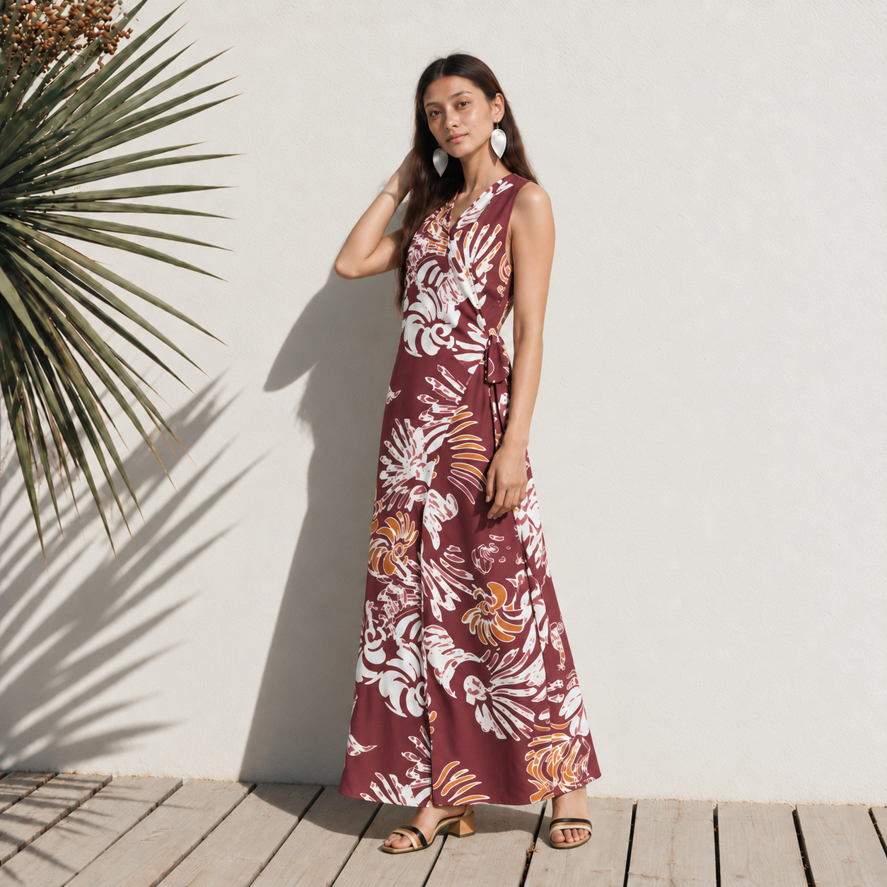 A woman stands on a wooden infront of a white wall wearing a sleeveless, wrap-style maxi dress with a side tie and a light maroon reef pattern.