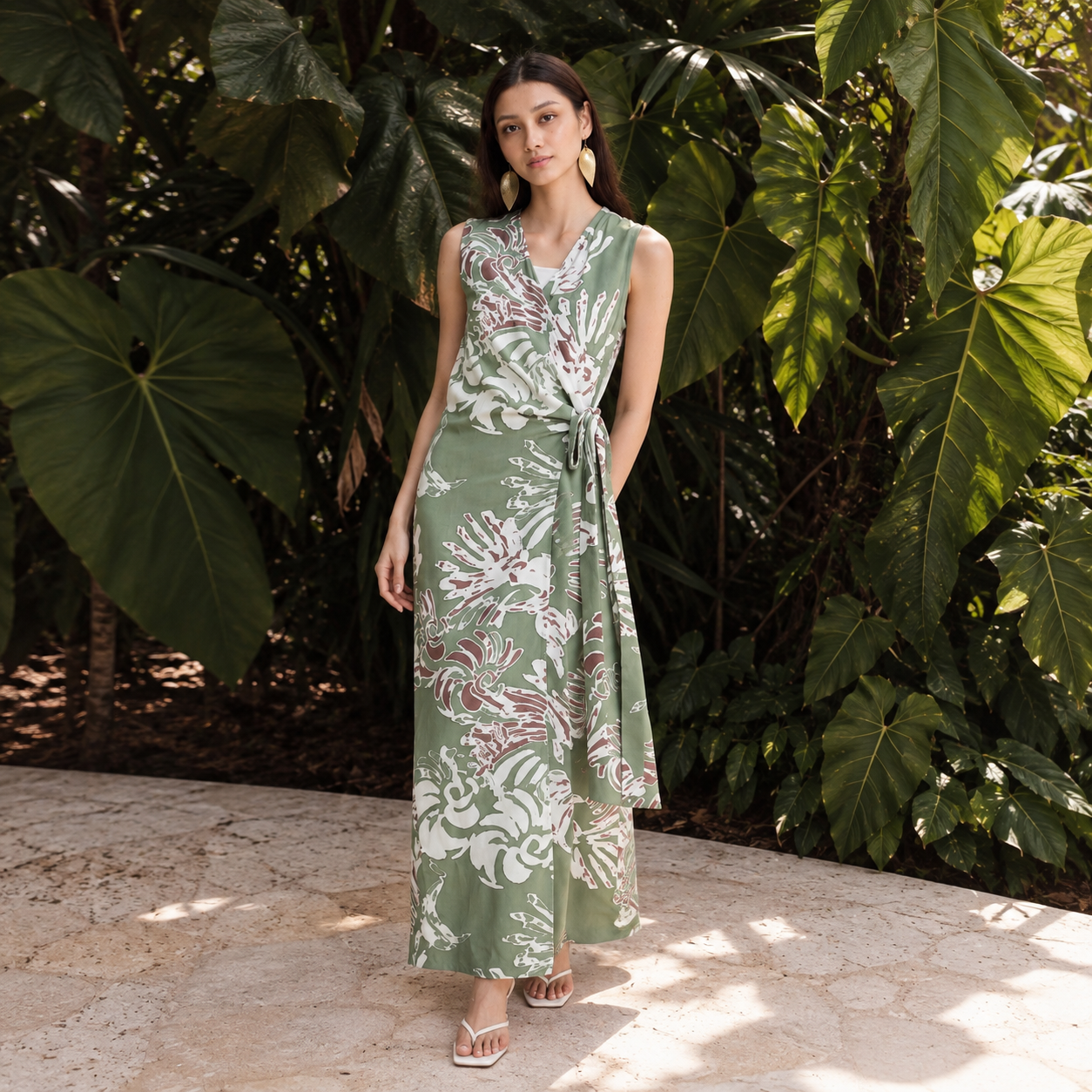 A woman stands by greenery wearing a sleeveless, wrap-style maxi dress with a side tie and a light green leaf pattern.