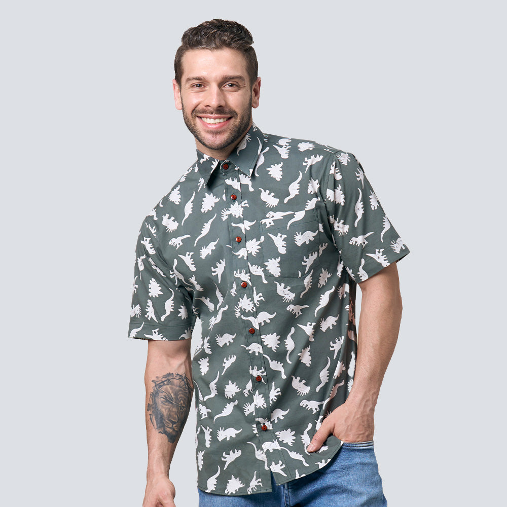 a man standing in front of a white wall wearing a batik shirt in olive dinosaurs pattern