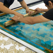 Two people pressing a large screen to print a blue and gold wave pattern onto a white sheet of material, likely silk or paper.