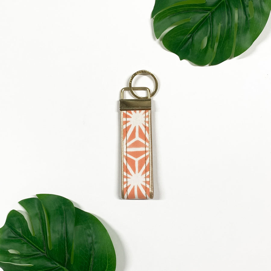 a lifestyle photo of a keyfob made of batik in the pattern peach firework against a neutral background surrounded by two tropical leaves