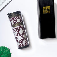 a lifestyle picture of a tumbler made of batik in the pattern plum firework with a black box from batik boutique, a tropical leaf and a book as decorations
