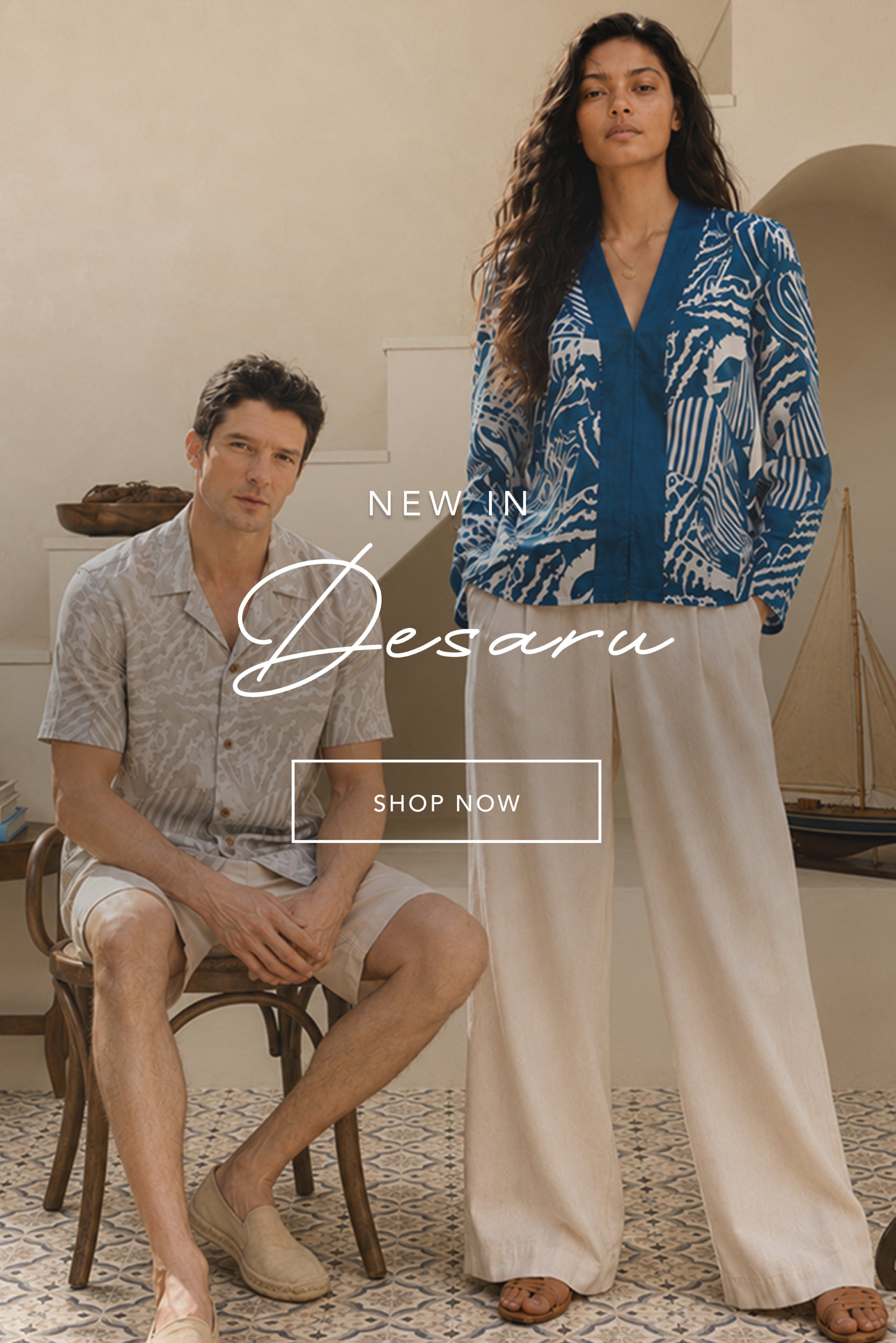 A man sits on a wooden chair wearing a light gray patterned short-sleeve shirt and beige shorts, while a woman stands beside him wearing a blue and white patterned cardigan and cream wide-leg pants. The image has a warm neutral interior background with text overlay reading 'NEW IN Desaru' and a 'SHOP NOW' button.