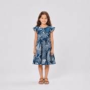 A young girl with long brown hair stands against a light gray studio background wearing a navy blue and white palm leaf print dress with ruffled cap sleeves and tan sandals.