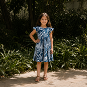 A young girl stands outdoors on a stone path wearing a blue and white leaf-patterned dress with ruffled cap sleeves and brown sandals, surrounded by lush green plants and trees.