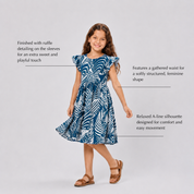 A young girl modeling a blue and white tropical print dress with text callouts describing the dress features against a light gray studio background.