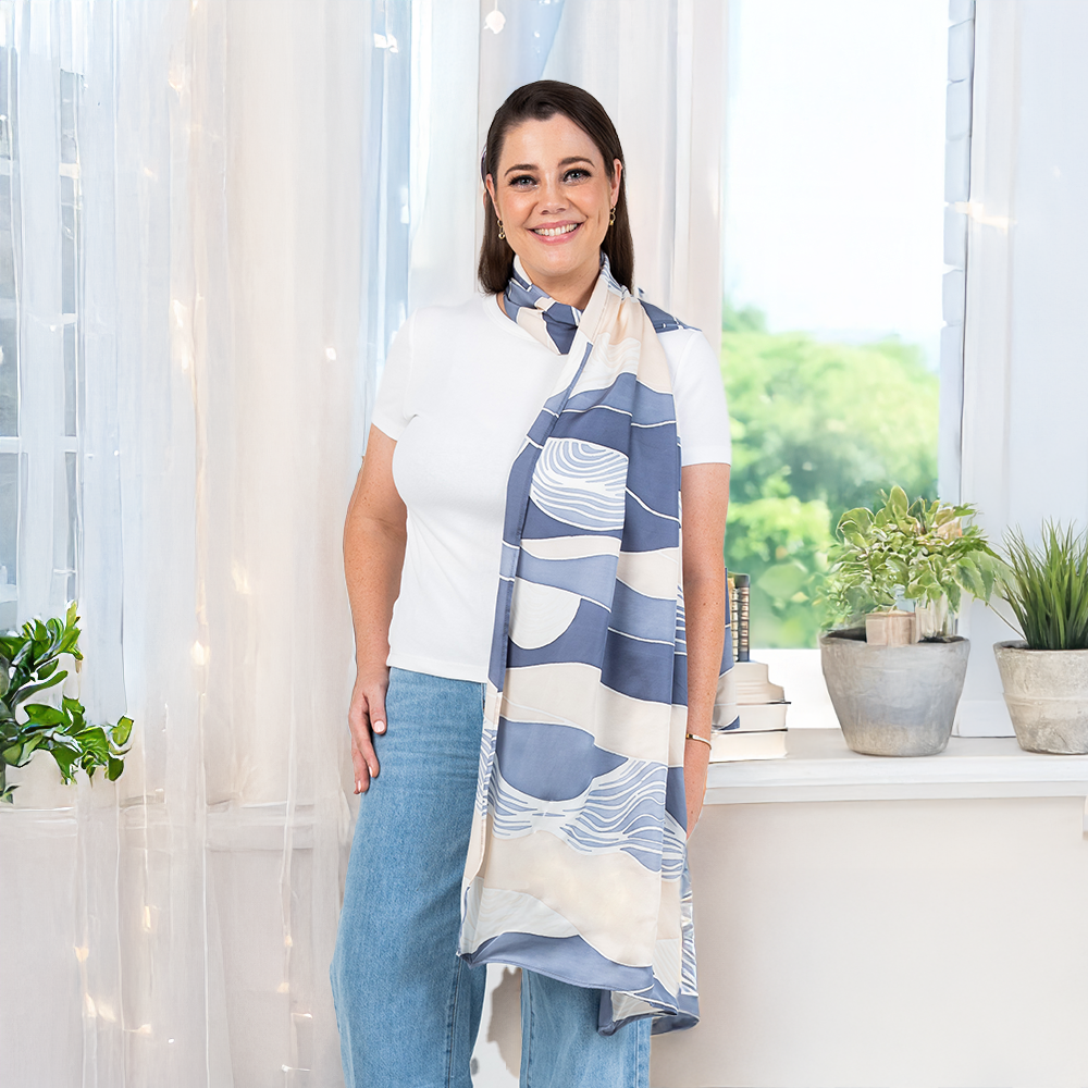 Woman wearing a white top and jeans, styled with a lavender and cream batik scarf featuring Bukit hill-inspired patterns, standing by a bright window with green plants and soft natural light.