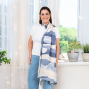 Woman wearing a white top and jeans, styled with a lavender and cream batik scarf featuring Bukit hill-inspired patterns, standing by a bright window with green plants and soft natural light.