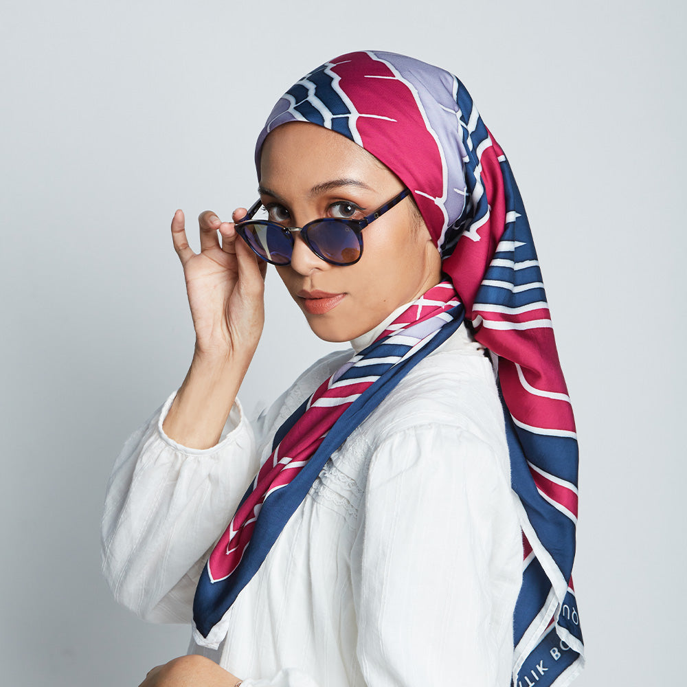 A women wearing sunglasses posing in front of white background wearing batik scarf in pattern fuchsia jambu