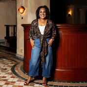 Woman standing in a stylish outfit with a patterned top and jeans in an indoor setting.