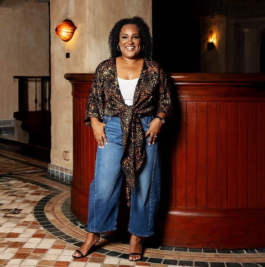 Woman standing in a stylish outfit with a patterned top and jeans in an indoor setting.