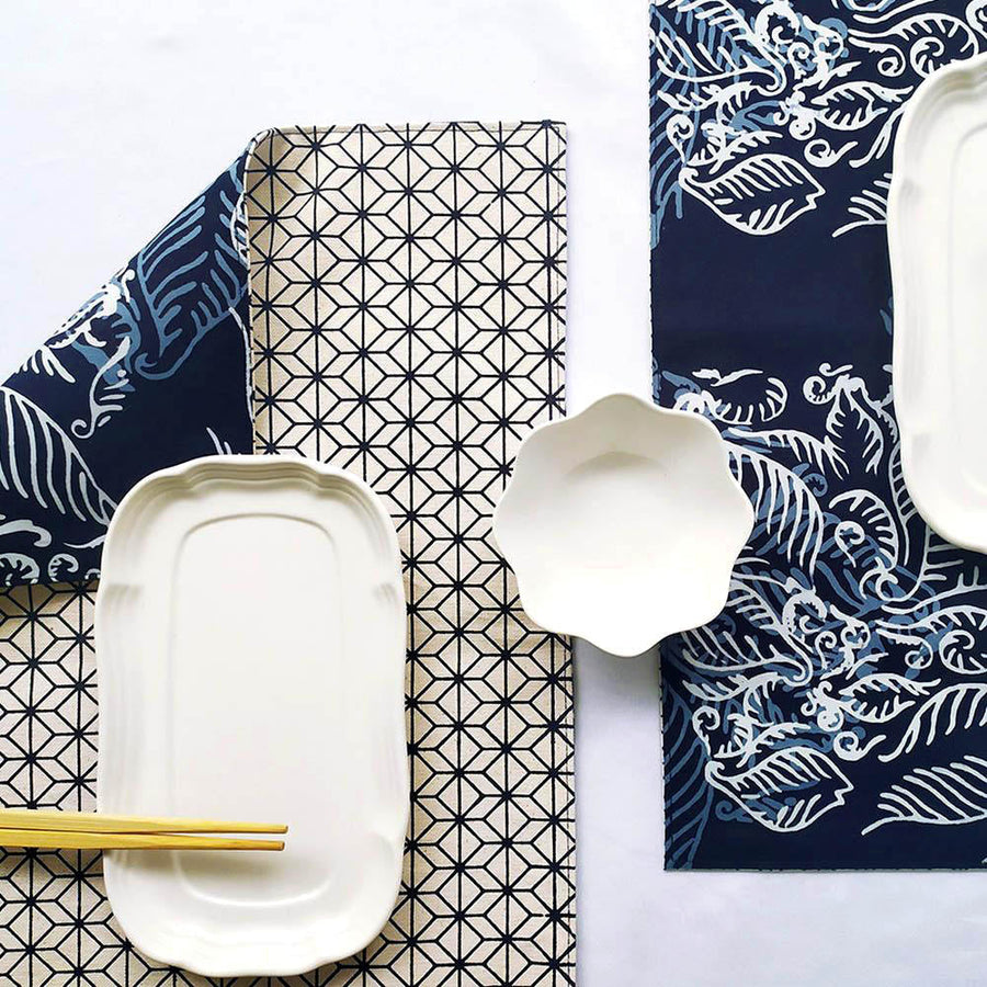 a lifestyle photo of reversible batik placemat in blue nautical fern pattern on dining table complete with plates and cutlery. the placemat showing front and back side of it.