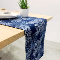 A batik table runner in blue shade color are display on dining table. The pattern of table runner is blue nautical fern