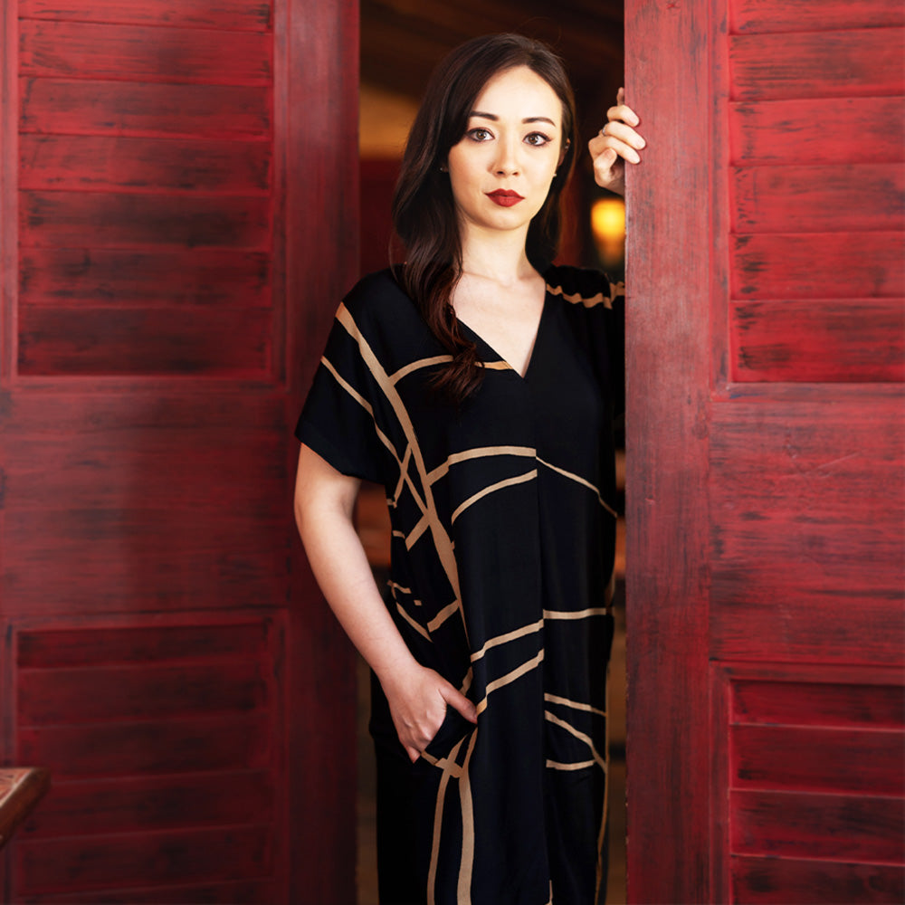 a lifestyle photo of a model posing in a batik dress in the pattern black ecru with red wooden doors beside her