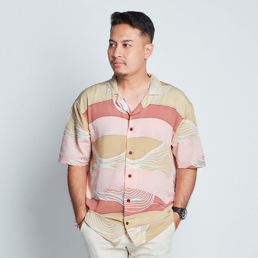 A man wearing batik cuban shirt in pink and neutral color combo. He is facing front with white wall at the back