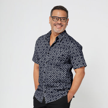 A man standing in front of white backdrop wearing black & grey batik motif in Arabesque. The model is facing side